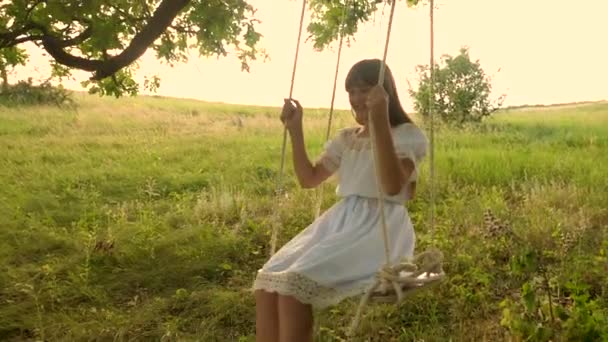 Teenage girl with long hair in a white dress laughs rolling on a swing under a summer oak tree in a warm season. Slow motion. — Stock Video