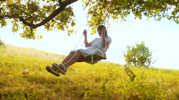 Jong meisje met lang haar swingt op een schommel onder een eikenboom. — Stockvideo