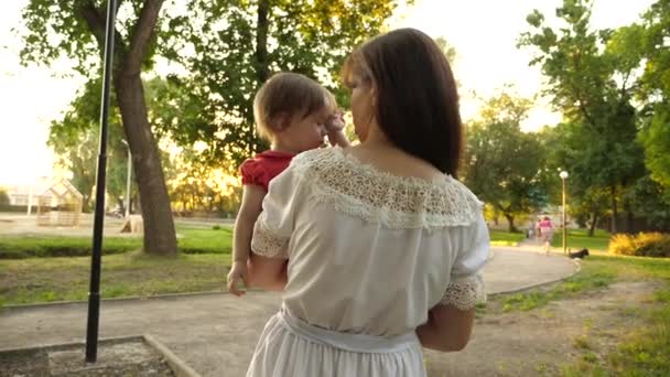 Criança esfrega os olhos enquanto se senta nos braços da mãe e caminha no parque no verão. Movimento lento — Vídeo de Stock