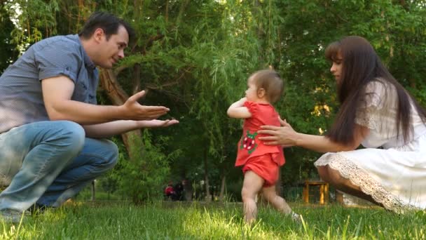 Малыш делает первые шаги, играя со смеющимися счастливыми родителями на газоне в городском парке. Медленное движение — стоковое видео
