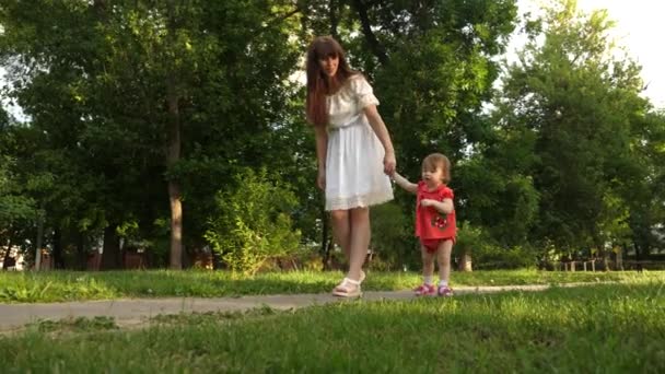 Baby macht erste Schritte, hält seine Mutter an der Hand und geht im Park spazieren. Mutter lacht über kleine Tochter. — Stockvideo