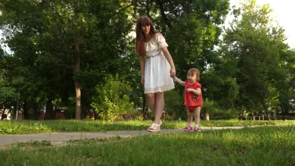 El bebé da los primeros pasos cogiendo a su madre de la mano y caminando en el parque. Mamá ríe caminando hija pequeña . — Vídeo de stock