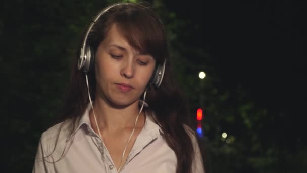 Chica en auriculares incluye canción en el teléfono táctil agitando su pelo largo y bailando sonriendo y kayfuya de la música por la noche en el parque de la ciudad. Primer plano — Vídeo de stock