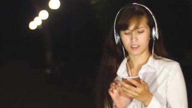 Kadın şarkı dinlerken kulaklık dans ediyor ve şehir gece park dokunmatik telefon aracılığıyla leafing söylüyor. Ağır çekim