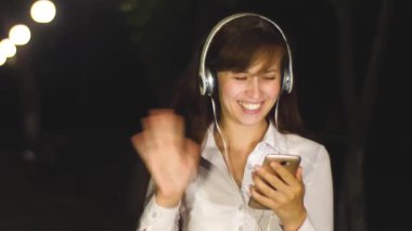 Kulaklık kız gece içinde değmek telefon el, gülüyor ve gülümsüyor şehir parkı ile iletişim kurar