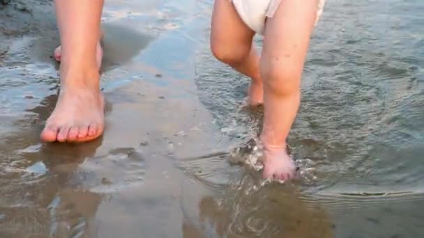 Bebê dá os primeiros passos ao longo da margem do rio, salpicando água nas laterais e andando junto com sua mãe. Pernas. Close-up. Motio lento — Vídeo de Stock