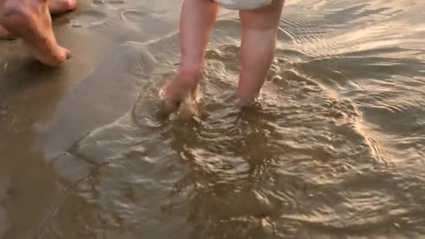 El bebé da los primeros pasos a lo largo de la orilla del río, salpicando agua en los lados y caminando junto con su madre. Piernas. Primer plano. Motio lento — Vídeo de stock