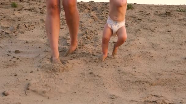 Μικρό μωρό στην πάνα κάνει τα πρώτα του βήματα περπατώντας κατά μήκος της άμμου στην παραλία κρατώντας το χέρι του με τη μητέρα του. Πόδια. Γκρο πλαν. Αργή κίνηση — Αρχείο Βίντεο