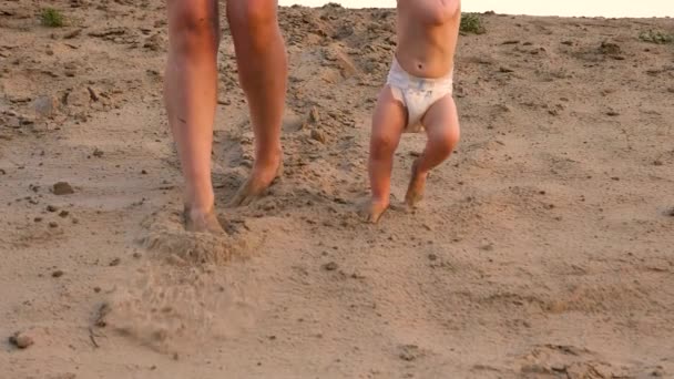 Liten bebis i blöja gör sin första stegen går längs sand på stranden håller hans hand med sin mor. Ben. Närbild. Slow motion — Stockvideo