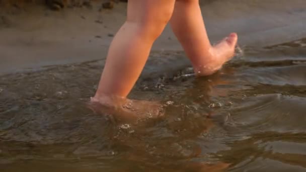 Pequeno bebê em fralda dá seus primeiros passos andando ao longo da margem do rio. Pernas. Close-up. Movimento lento — Vídeo de Stock