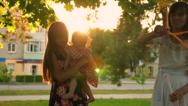 Мама и ее дочери ловят большие и прозрачные мыльные пузыри в городском парке и смеются в лучах солнца — стоковое видео