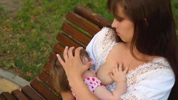 Bebé pequeno chupa leite do peito da mãe. Mãe está amamentando criança sentado no banco do parque — Vídeo de Stock