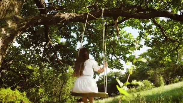 Hermosa chica con el pelo largo en vestido blanco, balanceándose en columpio en un parque de verano. Movimiento lento . — Vídeos de Stock