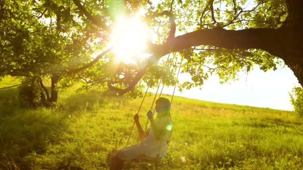 Glad tjej med långt hår, svängande på swing i ljusa strålar gyllene solnedgång och leende. Slow motion — Stockvideo