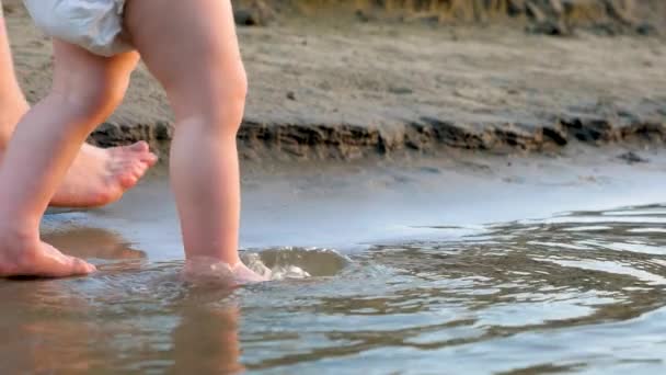 Bambino in pannolino fa i primi passi lungo la riva del fiume, spruzzando acqua intorno e camminando con sua madre. Gambe. Primo piano. Rallentatore — Video Stock