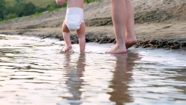 Criança de fralda vai com sua mãe ao longo da margem do rio. Família feliz andando na praia. Movimento lento . — Vídeo de Stock