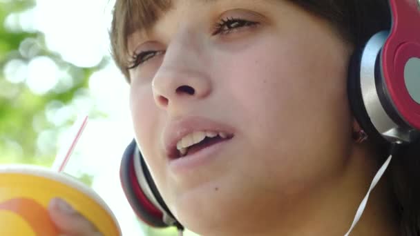 Menina em fones de ouvido bebe coquetel legal de tubo apreciando música de alto-falantes, risos e sorrisos. Close-up — Vídeo de Stock