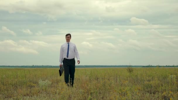 Üzletember, fehér ing és nyakkendő a kezében, háttérben a sötét felhők séta mező a táska segítségével — Stock videók