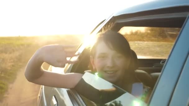 Piękna dziewczyna jeździ w samochodzie z rękami w oknie i uśmiecha się machając jej rękę w jasne światło Złotego słońca. Zwolnionym tempie. — Wideo stockowe