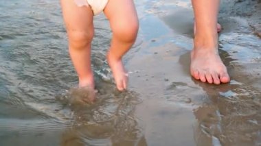 Bebek ilk adımları nehir banka, taraf su sıçramasına ve annesi ile birlikte yürüme alır. Bacaklar. Yakın çekim. Yavaş motio