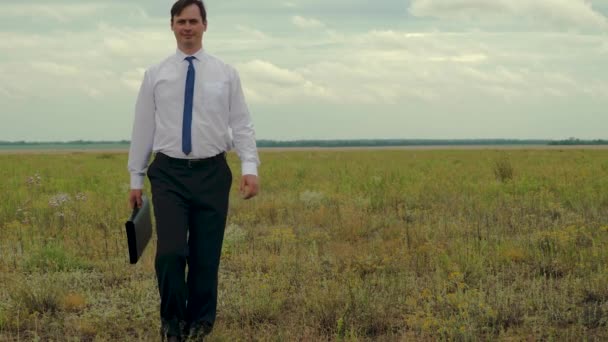 Business man in tie with portfolio walks across field and smiles. Slow motion — Stock Video