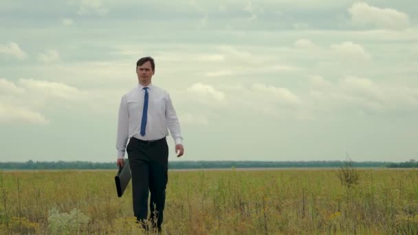 Üzletember, fehér ing és nyakkendő a kezében, háttérben a sötét felhők séta mező a táska segítségével — Stock videók