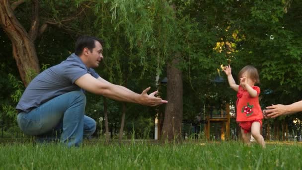 El pequeño bebé da los primeros pasos, jugando con los padres felices riendo en la hierba del césped en el parque de la ciudad. Movimiento lento — Vídeos de Stock