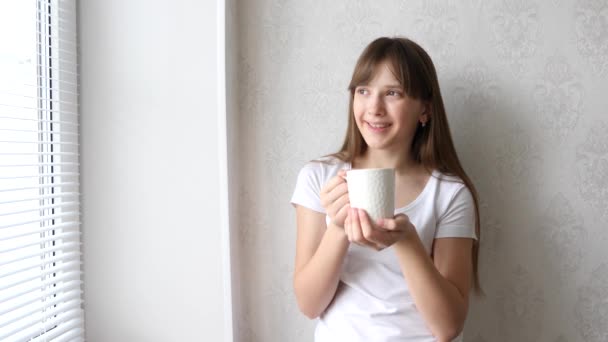Adolescente chica bebe té caliente en ventana y sonríe . — Vídeo de stock