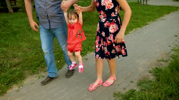 A mãe e o pai andam por aí de mãos dadas com um bebé pequeno, feliz e sorridente. A família está brincando com a criança andando pela estrada no parque da cidade. Movimento lento — Vídeo de Stock