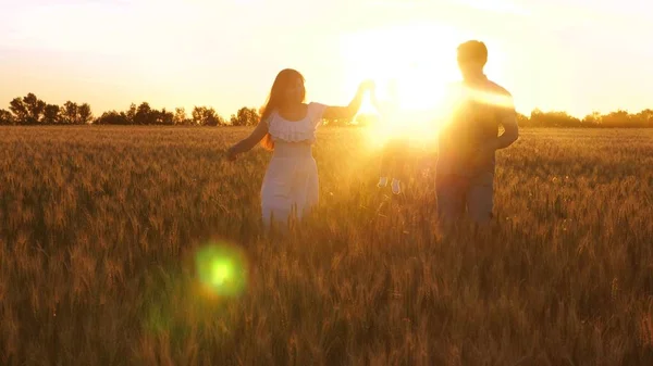 Mamãe e papai carregam a filha pequena nos braços através do campo de trigo em belos raios de pôr do sol. Bebê com os pais brincando e sorrindo em um campo com trigo . — Fotografia de Stock