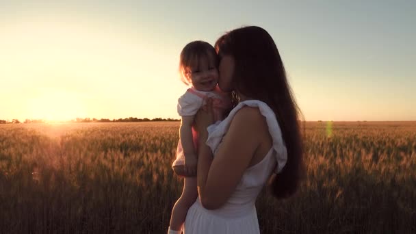 Mamá lleva a la pequeña hija en brazos a través del campo de trigo en hermosos rayos de la puesta del sol — Vídeo de stock