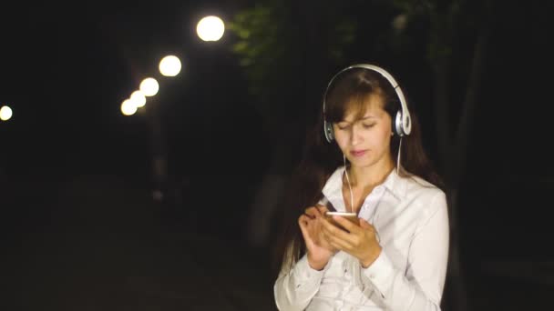 Woman in headphones listening to song dances and sings leafing through touch phone in city night park. Slow motion — Stock Video
