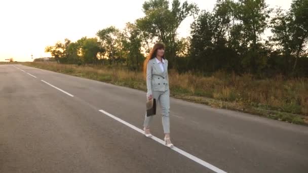 Mujer de negocios con maletín negro camina en traje ligero y zapatos de tacón alto blancos va fuera de la ciudad a lo largo de asfalto con marcas blancas, vista desde la parte delantera — Vídeos de Stock