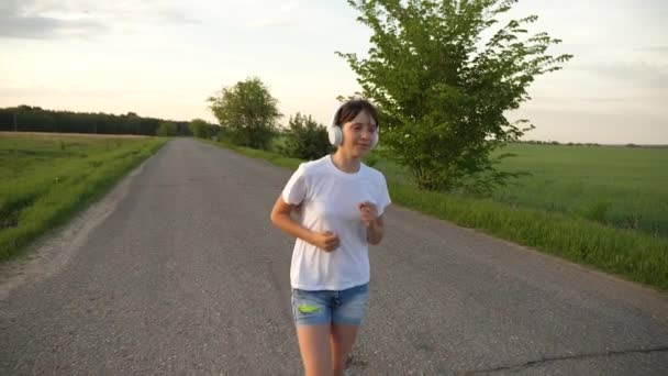 Hermosa chica haciendo ejercicios deportivos. chica en los auriculares de música corre a lo largo de la carretera. Movimiento lento — Vídeos de Stock