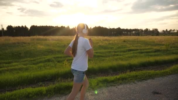 Chica joven entrena trotar, chica deportiva corre a lo largo del asfalto en el sol y escucha música en los auriculares. Movimiento lento — Vídeo de stock