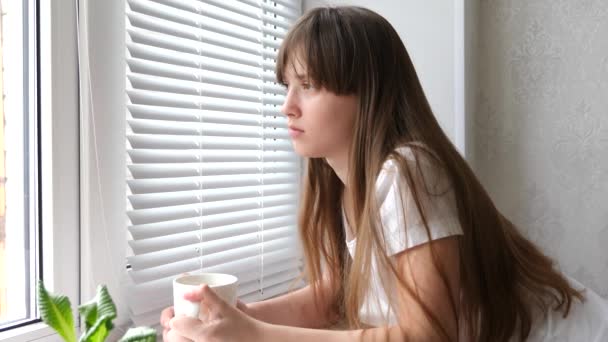 Chica somnolienta beber té caliente con vidrio blanco, chica triste mirando por la ventana — Vídeos de Stock