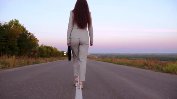 Mujer de negocios lleva maletín negro en la mano, mujer camina a lo largo de asfalto fuera de la ciudad con pantalones y chaqueta y zapatos blancos de tacón alto, una vista trasera — Vídeos de Stock