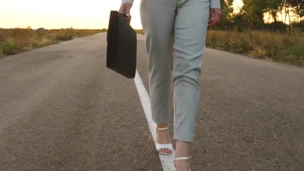 Mujer camina a lo largo de asfalto, fuera de la ciudad en pantalones y una chaqueta y en zapatos blancos de tacón alto, mujer de negocios lleva un maletín negro en la mano — Vídeo de stock
