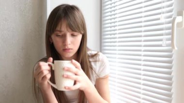 Çay içme ve pencere kenarında oturan üzgün kız. kız beyaz cam elinde tutan ve pencereden görünüyor.