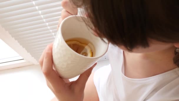 Menina bonita senta-se no peitoril da janela perto da janela e bebe chá com limão. Prevenção de resfriados. chá de vitamina contra resfriados — Vídeo de Stock