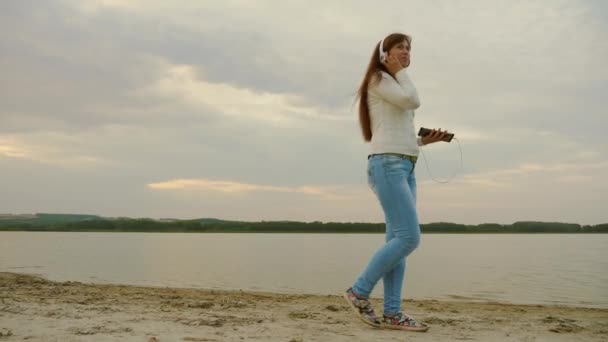 戴耳机的女孩晚上在海滩上用平板电脑散步, 听音乐. — 图库视频影像