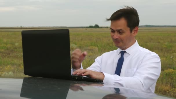 Happy businessman in blue and white shirt works in field on computer on the roof of car. — Stock Video