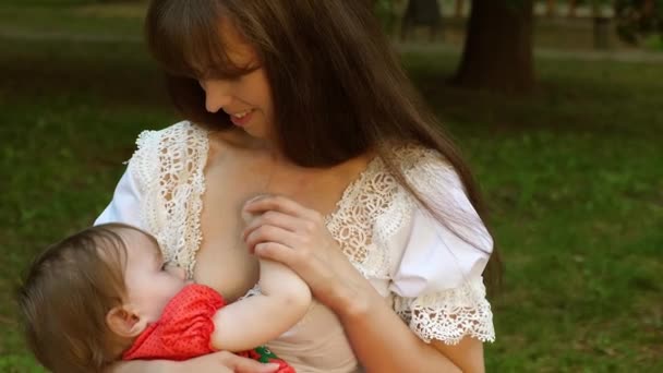 Small baby sucks milk from his mom breast. Mom is breastfeeding child sitting on park bench and smiling — Stock Video