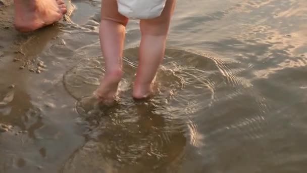 Мама веде маленьку дитину в підгузниках вручну на пляжі. дитячі ноги ходять по чистій воді на пляжі. Повільний рух — стокове відео
