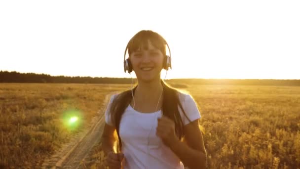 Спортивная девушка в наушниках занимается бегом трусцой, молодая девушка тренируется на закате и слушает музыку — стоковое видео