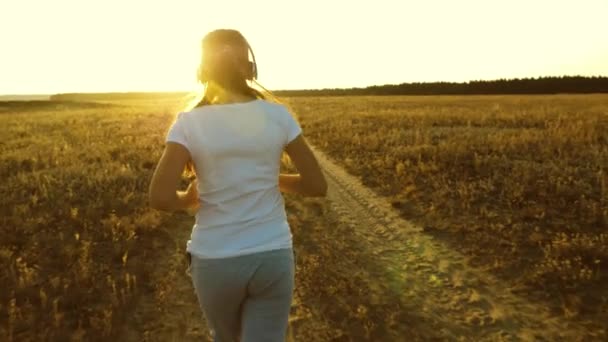 Sport flicka i hörlurar är engagerad i jogging, en ung flicka är utbildning vid solnedgången och lyssna på musik — Stockvideo