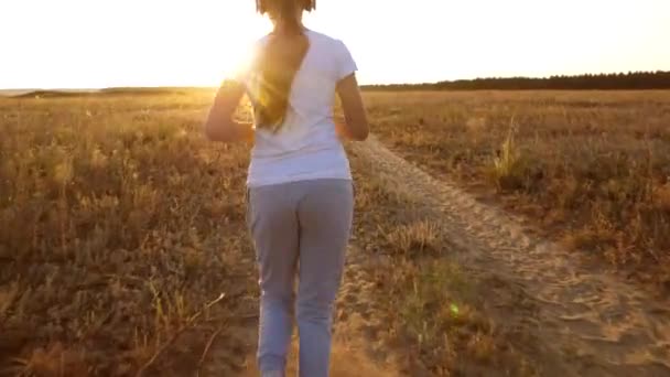 Ein junges Mädchen trainiert bei Sonnenuntergang und hört Musik, ein Sportmädchen mit Kopfhörern joggt. zurück — Stockvideo