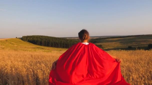 Mädchen spielt Superheldin, die mit Weizen in rotem Mantel über das Feld gegen den blauen Himmel läuft. Zeitlupe. — Stockvideo
