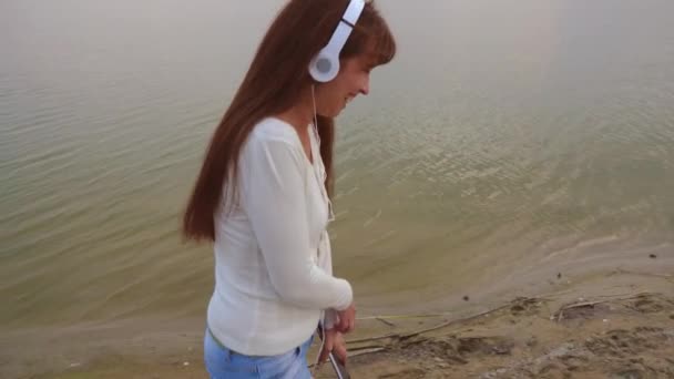 Flicka i hörlurar går på stranden med en tablett och lyssnar på musik, vacker flicka skrattar och ler — Stockvideo