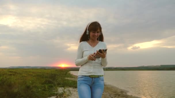 Dívka v paprscích slunce chůze po pláži a kontrola pošty na tabletu online. dívka s sluchátka, procházky po pláži s tabletem a poslechu hudby. — Stock video